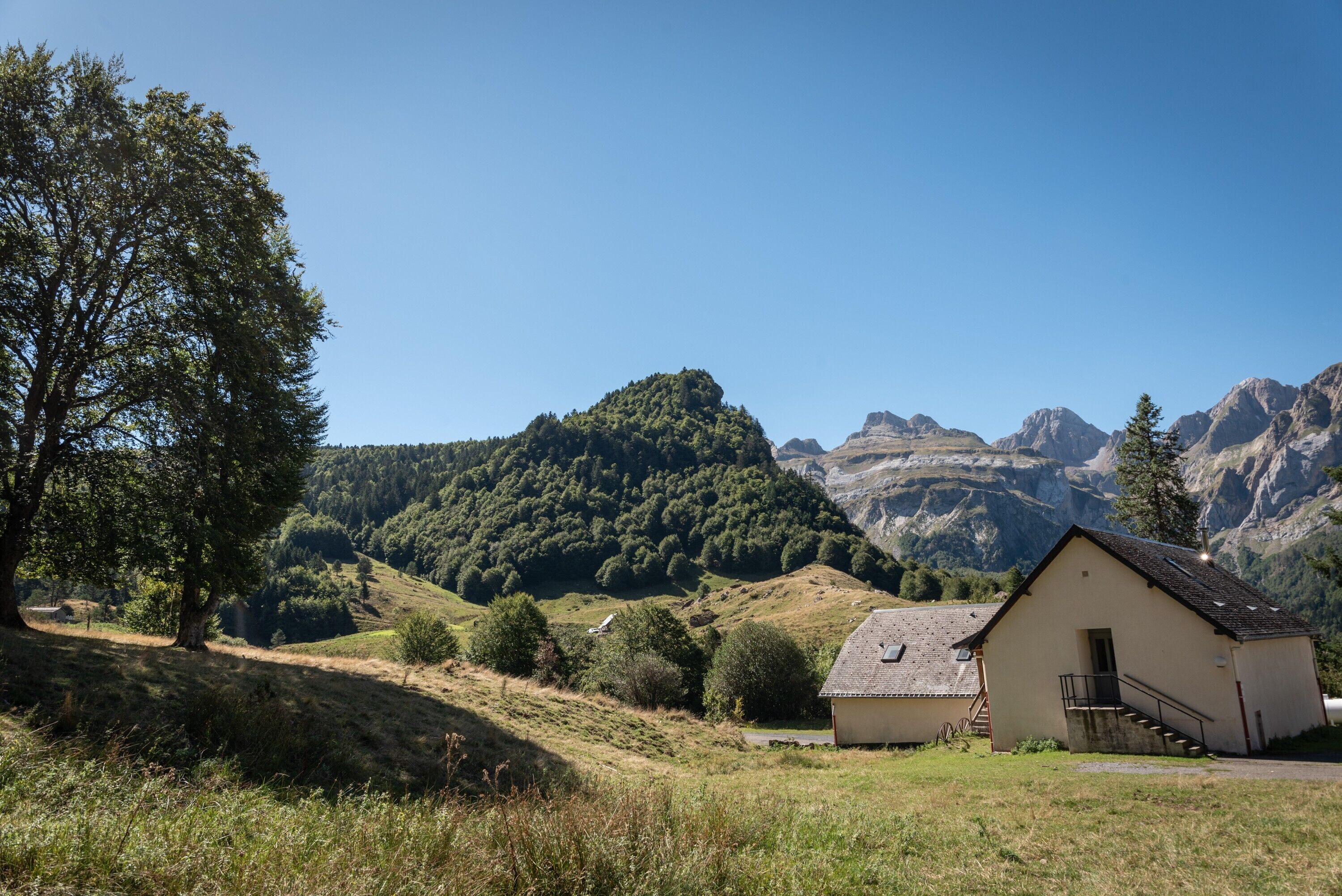 Un petit bâtiment doté d’un escalier, entouré d’une zone herbeuse et d’arbres, avec des montagnes en arrière-plan.