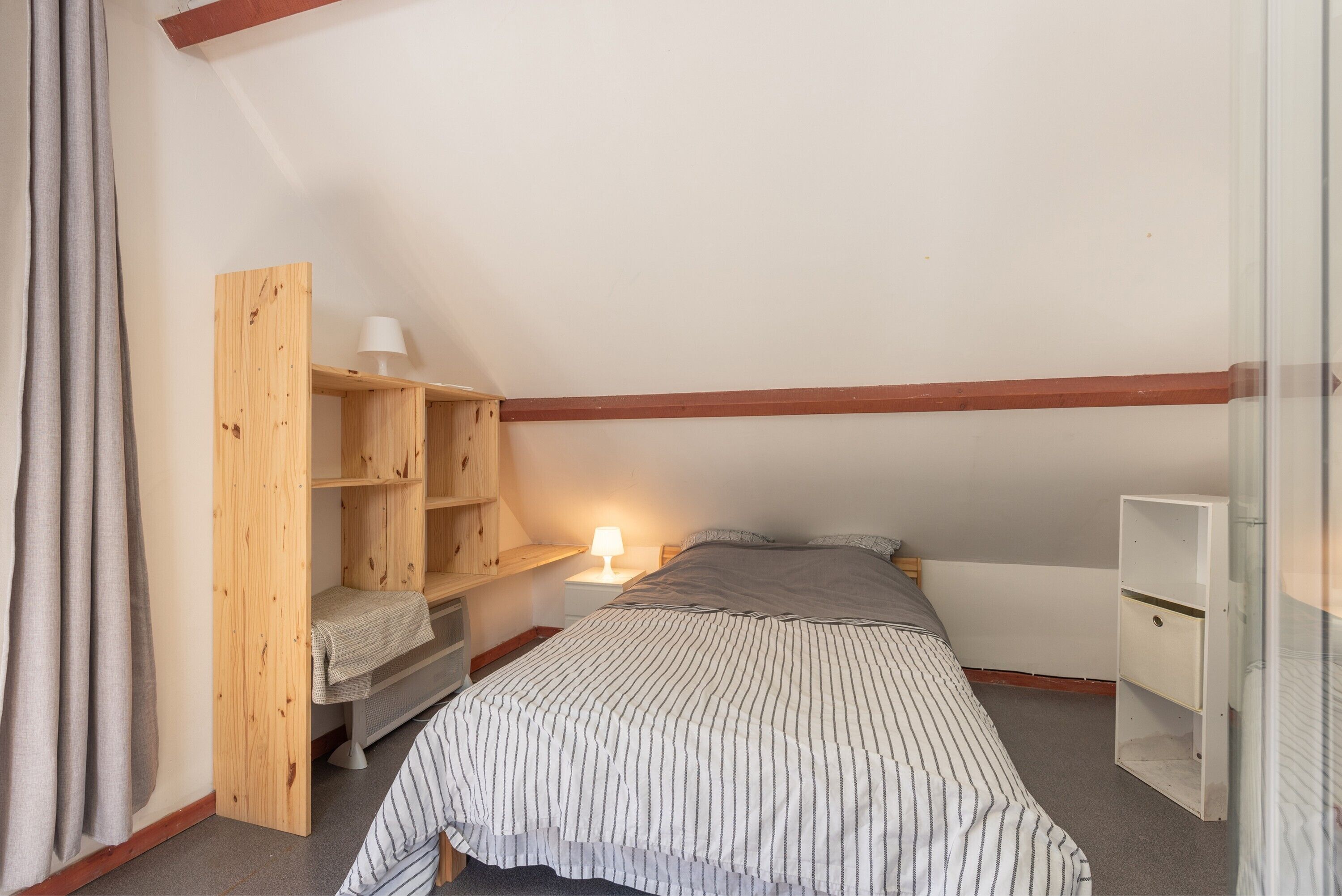 A small, single-bed room with a sloped ceiling, a wooden wardrobe, a bedside table with a lamp, and a window.