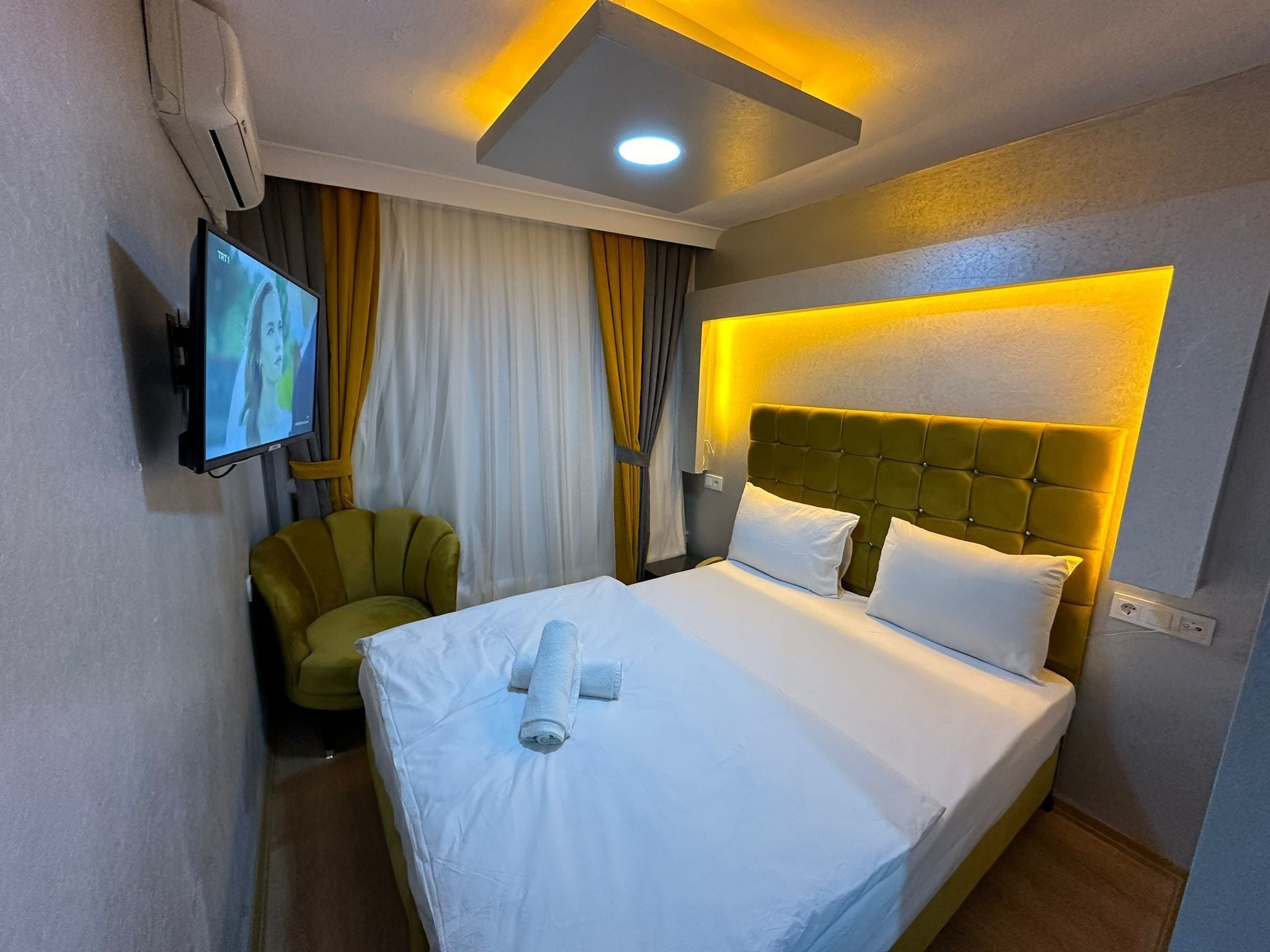Hotelkamer met een bed, een aan de muur bevestigde televisie, een groene fauteuil en een plafondlamp.