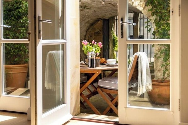 An open door leading to a dining area with a table set for two, a vase with flowers, and a view of a stone archway.