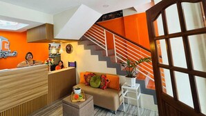 A hotel reception area with a wooden reception desk, a sofa with colorful cushions, a small table with a tray of flowers, and a staircase with orange accents. - OYO 1158 Kegama Residences 2 (Dasmariñas)