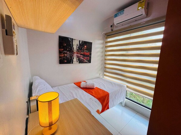 A small, modern bedroom with a bed, a nightstand, a lamp, and a window with blinds.