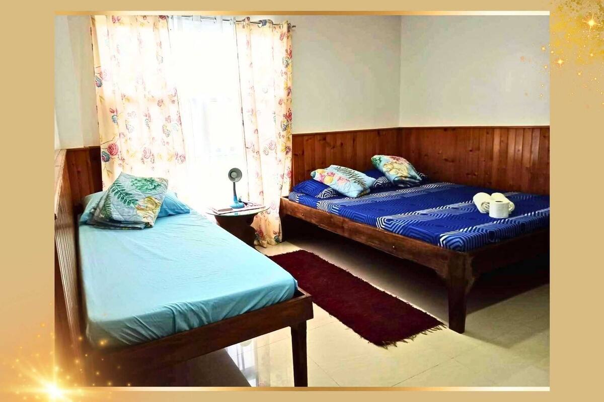 Une chambre avec deux lits simples, une tête de lit en bois, une petite table de chevet, une commode et une fenêtre agrémentée de rideaux à motifs floraux.