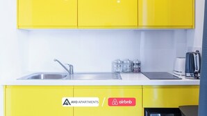 Eine Küche mit gelben Schränken, einer Spüle aus Edelstahl und einer Kaffeemaschine. Drei Glasbehälter sind mit „ZUCKER“, „TEE“ und „KAFFEE“ beschriftet. Die Küchenarbeitsplatte verfügt über ein schwarzes Induktionskochfeld und einen Toaster.