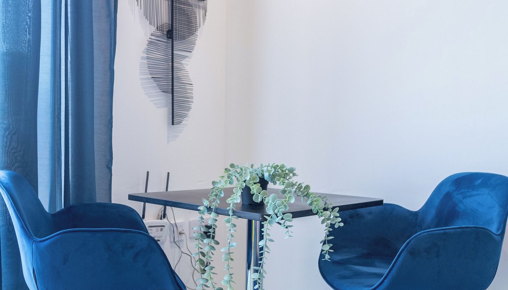 A modern living room with a dark wooden table, two blue chairs, a potted plant, and a decorative wall piece.