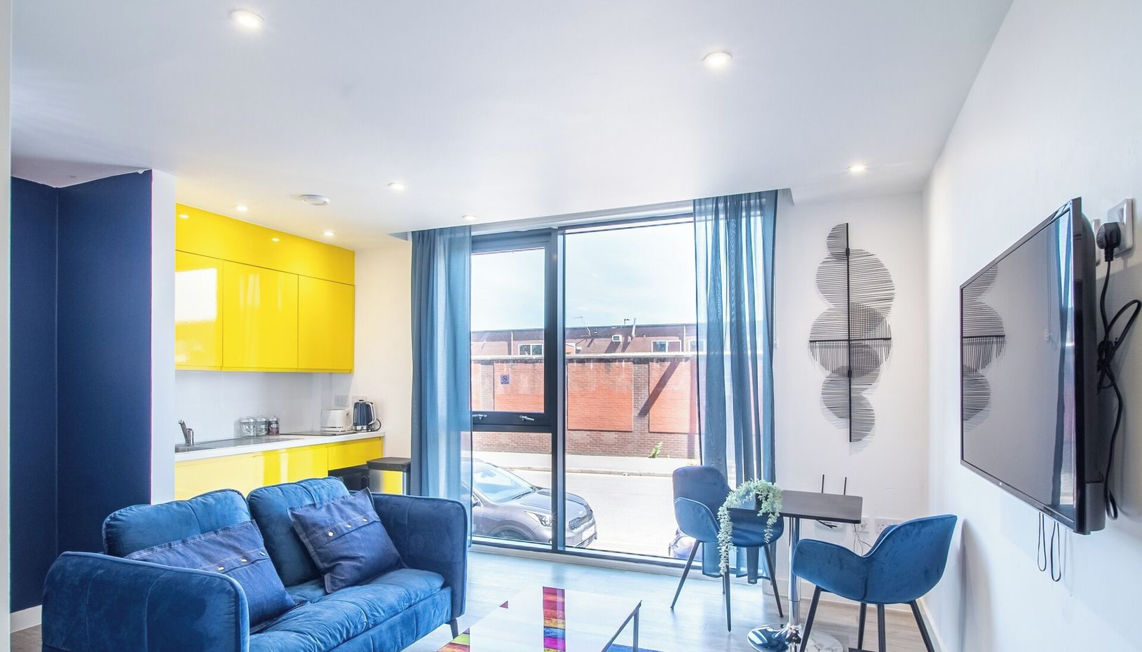 A modern apartment with a blue sofa, a dining table, and a kitchen with yellow cabinets.