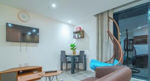 Ein modernes Wohnzimmer mit Fernseher, Esstisch mit Stühlen, einer Couch, einem kleinen Holzschrank und einem Hängesessel.