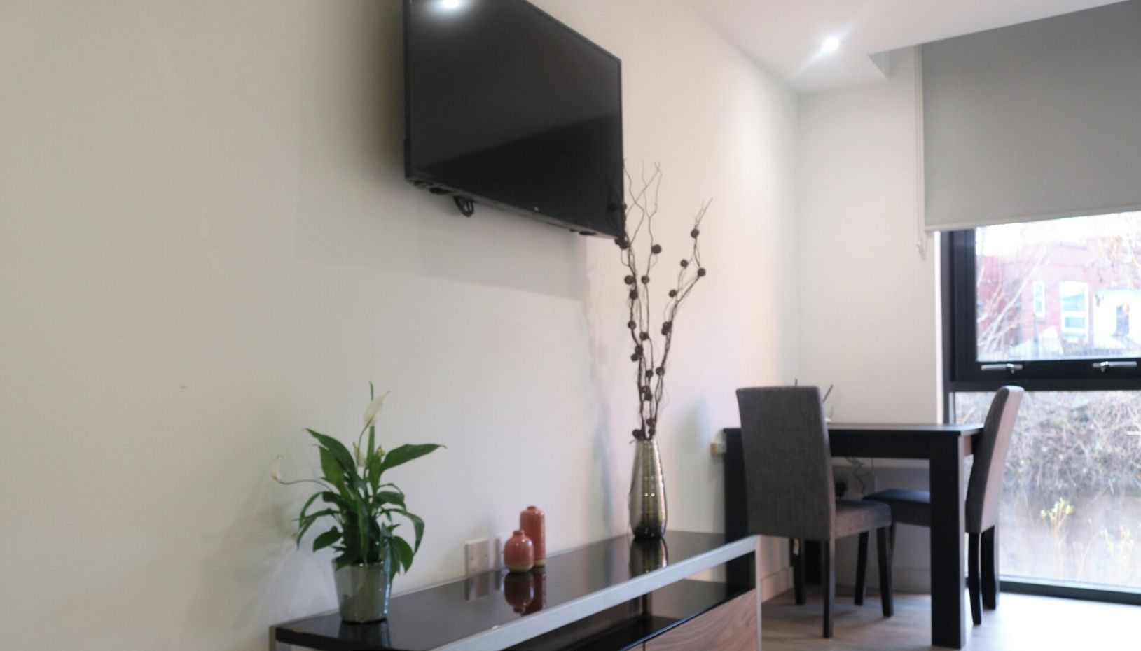 A modern living room with a flat-screen TV mounted on the wall, a dining area with a table and chairs, and a potted plant on a console table.