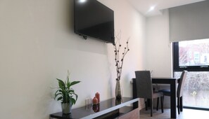 A modern living room with a flat-screen TV mounted on the wall, a dining area with a table and chairs, and a potted plant on a console table.