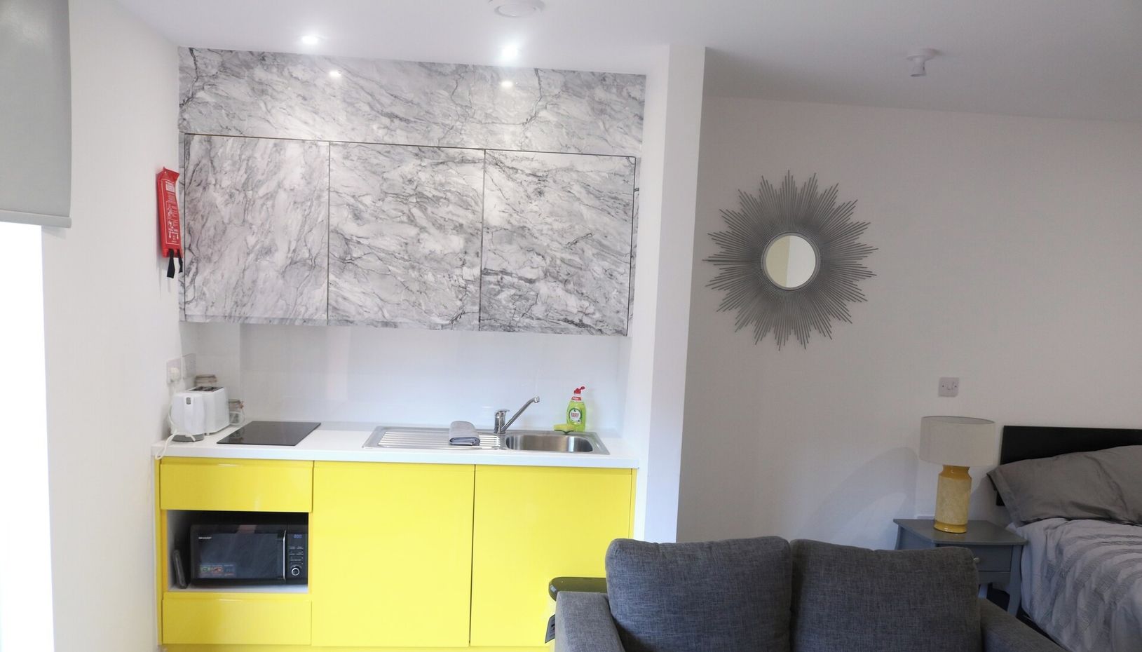 A compact living space with a kitchen area featuring a marble backsplash, a yellow cabinet, and a grey sofa.