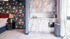 A compact room with a bed, a small bedside table, a nightstand, a wall with floral wallpaper, a marble backsplash, a sink, a kettle, and a trash bin. - Lovely Studio Sheffield Centre – APT 11 (Sheffield)