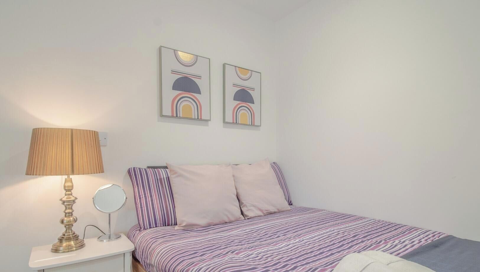 Un dormitorio con una cama, una mesita de noche con lámpara, un espejo y dos cuadros en la pared.