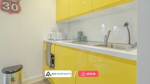 Una cocina con armarios amarillos, una encimera blanca, una cafetera, un hervidor y un lavabo con grifo. Hay un cubo de basura y un cartel en la pared.