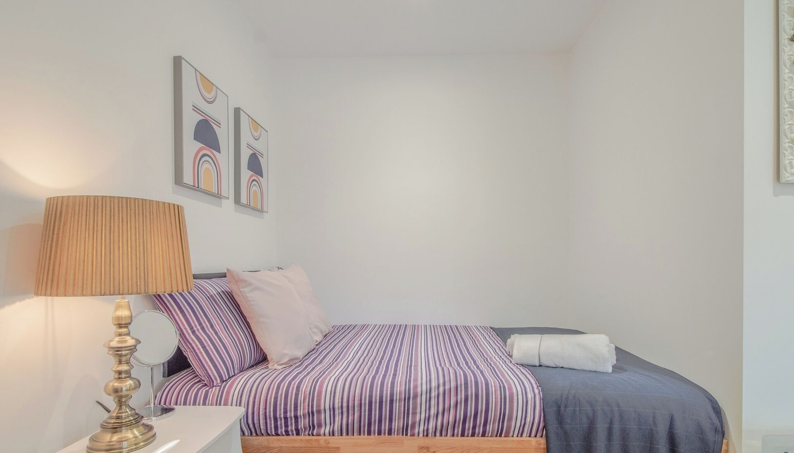 A bedroom with a bed, a bedside table with a lamp, and a dresser. There are two framed pictures on the wall.