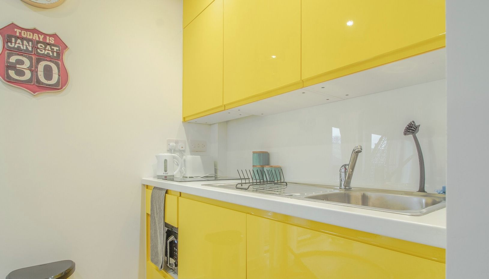 A kitchen with yellow cabinets, a white countertop, a coffee maker, a kettle, and a sink with a faucet. There is a trash bin and a sign on the wall.