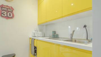 A kitchen with yellow cabinets, a white countertop, a coffee maker, a kettle, and a sink with a faucet. There is a trash bin and a sign on the wall.