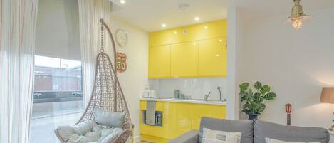 A modern apartment with a hanging chair, a sofa, a kitchen with yellow cabinets, and a visible clock.