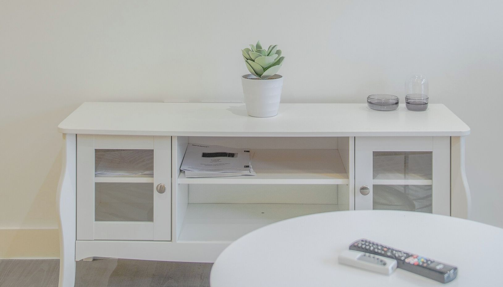 A white TV stand with a potted plant, two remote controls, and a small glass container.