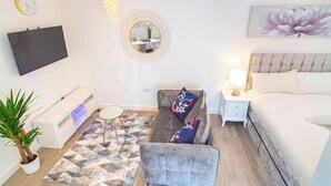 Una sala de estar moderna con sofá, sillón y cama. Hay un televisor montado en la pared, un espejo redondo y un cuadro sobre la cama.