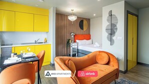 Un appartement moderne comprenant une cuisine, un coin repas et une chambre. La cuisine est dotée de meubles jaunes, d’une table à manger avec des chaises orange et d’un lustre. La chambre comprend un lit avec une tête de lit orange, une œuvre d’art murale et une fenêtre.