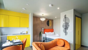 Un apartamento moderno con cocina, comedor y dormitorio. La cocina tiene armarios amarillos, una mesa de comedor con sillas naranjas y una lámpara de araña. El dormitorio cuenta con una cama con cabecera naranja, una obra de arte en la pared y una ventana.