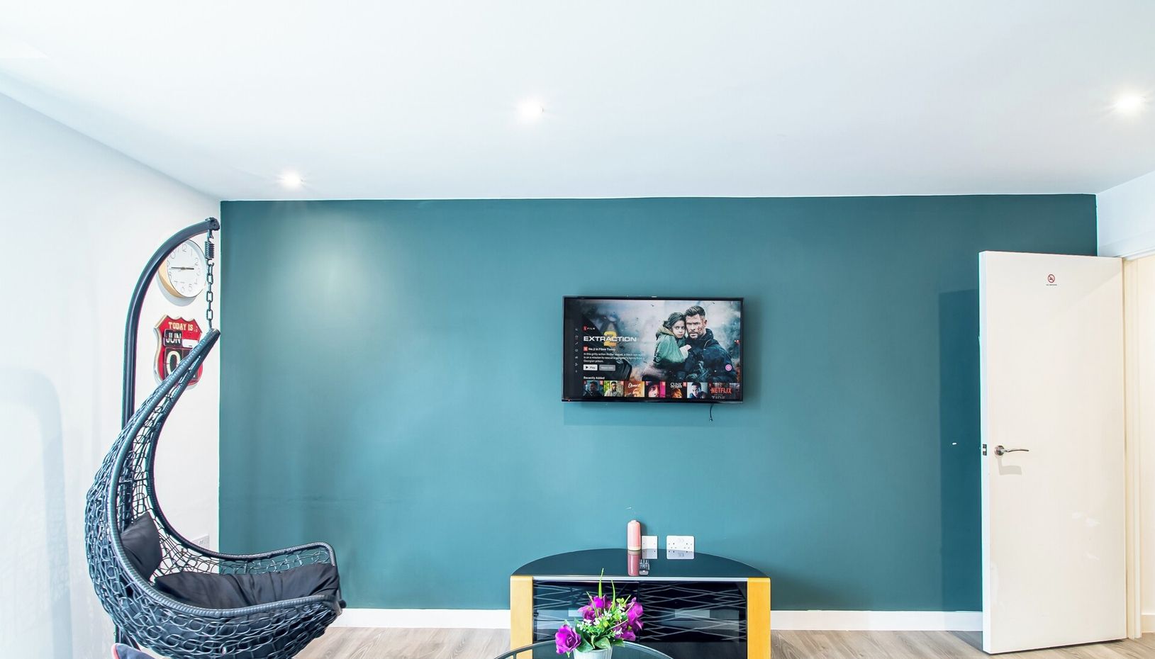 A modern living room with a teal accent wall, a mounted TV, a black coffee table with a vase of flowers, and a unique hanging chair.
