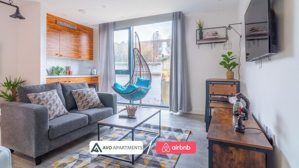 A modern living room with a grey sofa, a hanging chair, a wooden kitchen area, and a large window with a view. - Creative Studio With Rustic Charm – APT 22 (Sheffield)