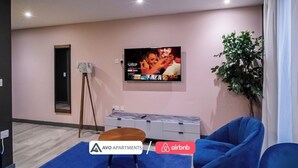 A modern living room with a flat-screen TV mounted on the wall, a standing lamp, a potted plant, and a blue sofa. - The Noir Apartment Sheffield – APT 12 (South Yorkshire)