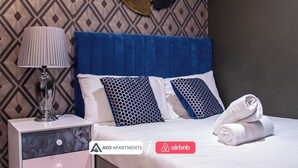 A neatly made bed with blue and patterned pillows, a bedside lamp, and a folded towel on a nightstand. The room features a patterned wallpaper and a dark wall. - The Noir Apartment Sheffield – APT 12 (South Yorkshire)