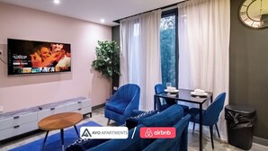 A modern living room with a flat-screen TV, blue armchairs, a dining table with white plates, and a potted plant. - The Noir Apartment Sheffield – APT 12 (South Yorkshire)