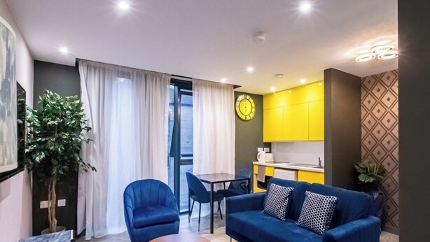 A compact living space with a dining area, kitchenette, and a large window with curtains. - The Noir Apartment Sheffield – APT 12 (South Yorkshire)