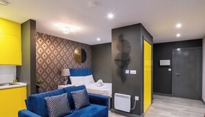 Een modern appartement met een blauwe bank, een bed met witte beddengoed, een behang met patroon en een gele keuken.