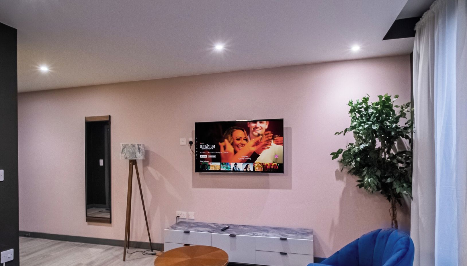 A modern living room with a flat-screen TV mounted on the wall, a standing lamp, a potted plant, and a blue sofa.