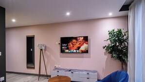 A modern living room with a flat-screen TV mounted on the wall, a standing lamp, a potted plant, and a blue sofa. - The Noir Apartment Sheffield – APT 12 (South Yorkshire)