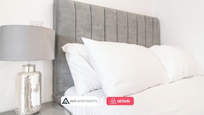A neatly made bed with a grey upholstered headboard, white pillows, and a metallic lamp on a bedside table.