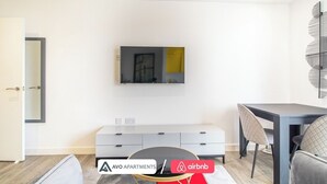 A modern living room with a flat-screen TV mounted on the wall, a grey sofa, a dining table with chairs, and a small cabinet below the TV.