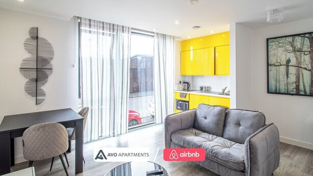 A modern apartment with a dining area, a grey sofa, a kitchen with yellow cabinets, and a large window with curtains.
