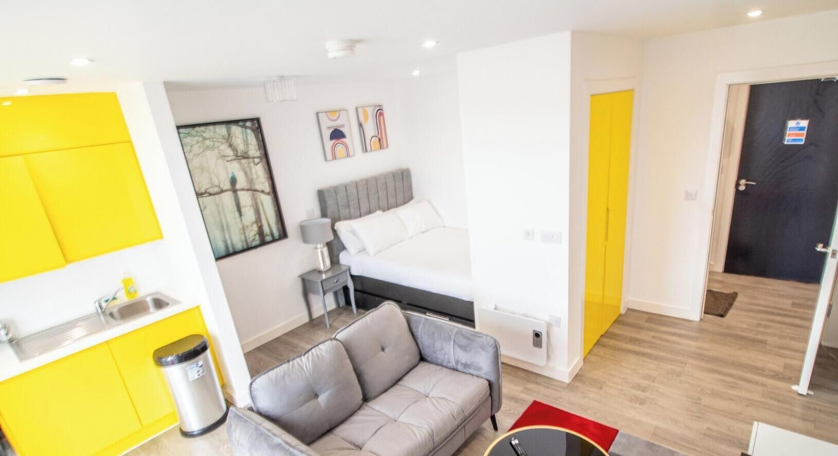 A modern apartment with a kitchenette, a grey sofa, a bed, and a small table.