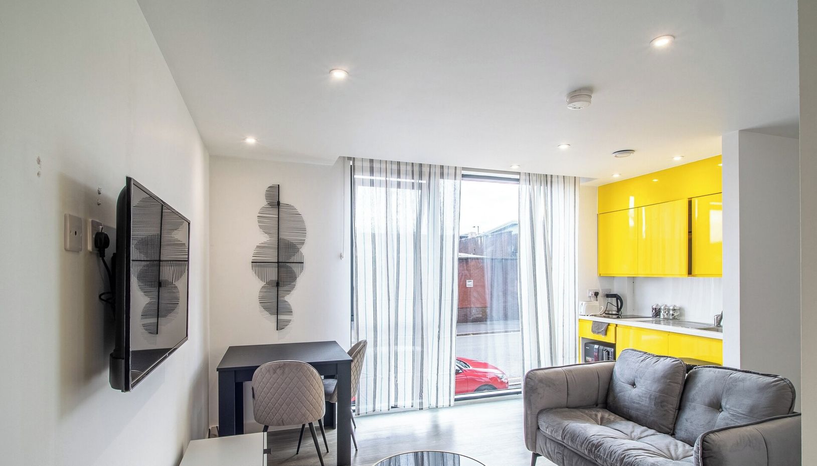 A modern living room with a grey sofa, a dining table with chairs, a black TV mounted on the wall, and a yellow kitchen in the background.