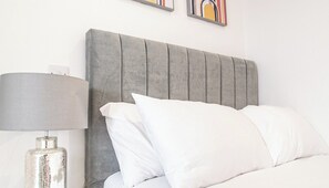 Una cama bien hecha con cabecera tapizada de gris, almohadas blancas y una lámpara metálica sobre una mesita de noche.
