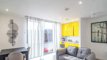 A modern apartment with a dining area, a grey sofa, a kitchen with yellow cabinets, and a large window with curtains.