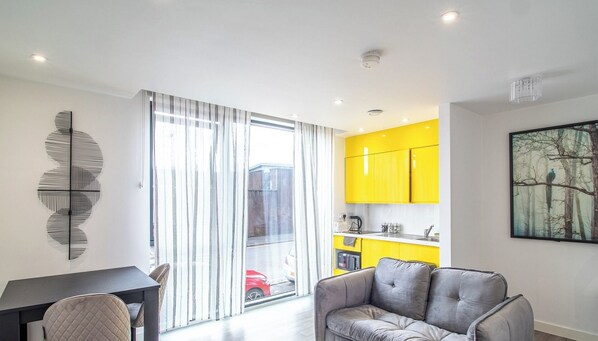 Un apartamento moderno con comedor, un sofá gris, una cocina con armarios amarillos y un amplio ventanal con cortinas.