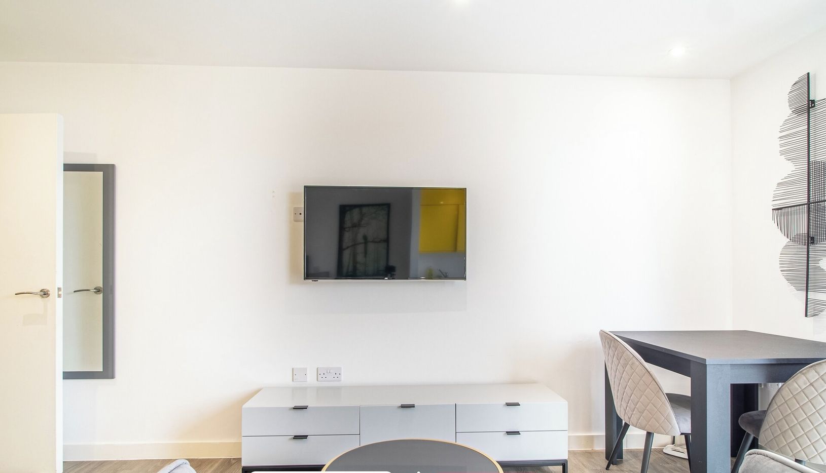 A modern living room with a flat-screen TV mounted on the wall, a grey sofa, a dining table with chairs, and a small cabinet below the TV.