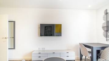 A modern living room with a flat-screen TV mounted on the wall, a grey sofa, a dining table with chairs, and a small cabinet below the TV.