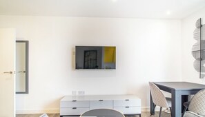Una sala moderna con un televisor de pantalla plana montado en la pared, un sofá gris, una mesa de comedor con sillas y un mueble bajo el televisor.