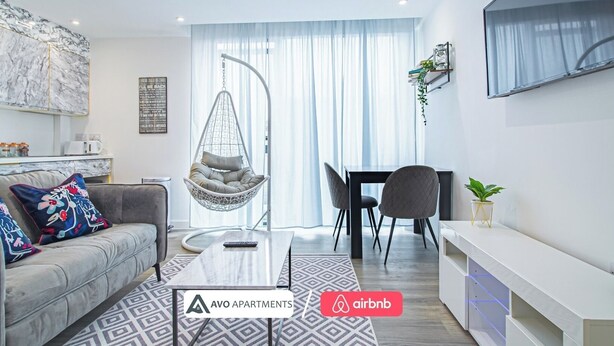 A modern living room with a sofa, hanging chair, dining table, and chairs. There is a TV mounted on the wall, a plant on a shelf, and a sign for AVO APARTMENTS and Airbnb. - Stylish Central Studio With Hanging Chair – APT 2 (Sheffield)