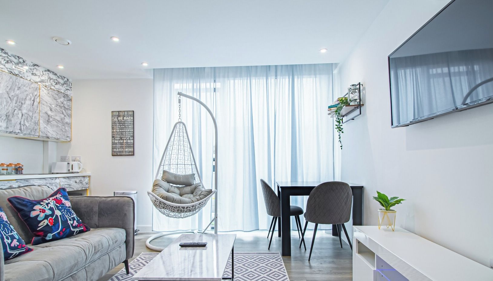 En moderne stue med sofa, hængestol, spisebord og stole. Der er et fjernsyn monteret på væggen, en plante på en hylde, og et skilt for AVO APARTMENTS og Airbnb.