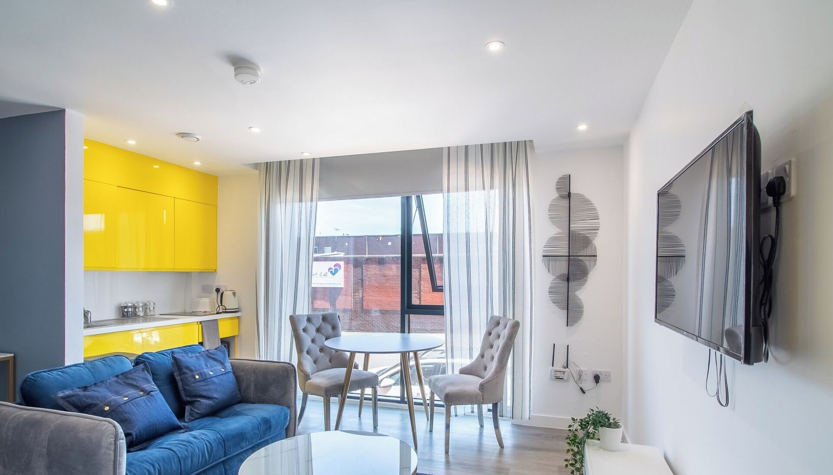 A modern apartment with a living area, dining space, and kitchen. It features a blue sofa, a round dining table with chairs, and a kitchen with yellow cabinets.