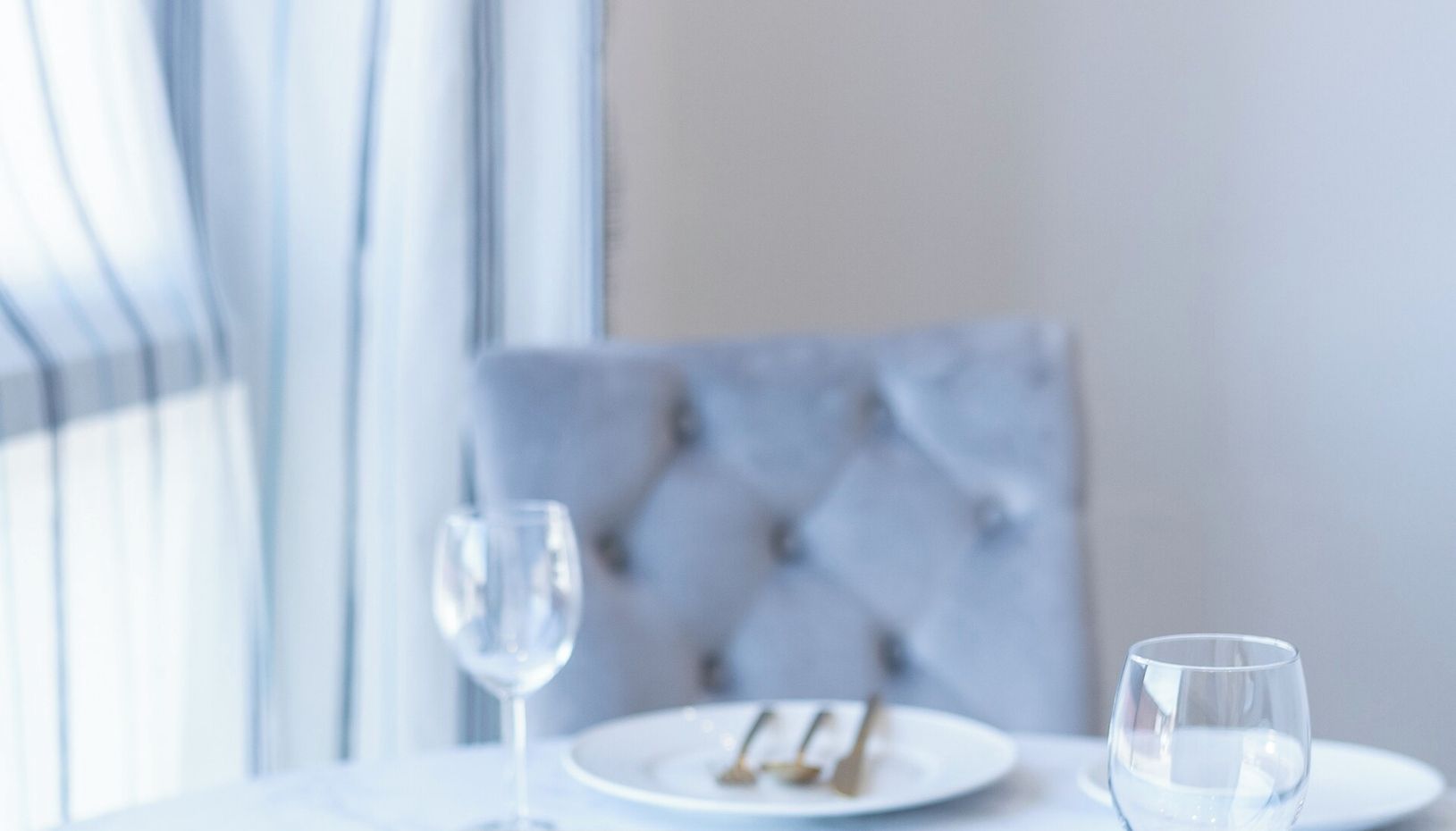 A dining table with two wine glasses and a plate of food, set against a modern interior with a tufted chair and a window with curtains.