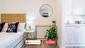 Une chambre à coucher avec une tête de lit capitonnée, une table de chevet avec une lampe, un miroir et une plante en pot. La pièce a un plancher en bois et un mur blanc.